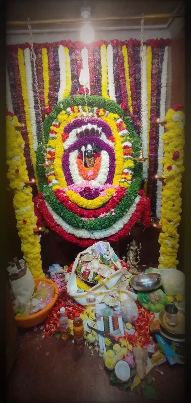 ಸುತ್ತುಗಳಾಗಿ ಹೂವಿನಿಂದ ಮತ್ತು ಉದ್ದದ ಹೂಮಾಲೆಗಳಿಂದ ಅಲಂಕರಿಸಿದ ಗುಡಿ; ಮುಂದೆ ಪೂಜಾ ಸಾಮಗ್ರಿ ಮತ್ತು ಕಾಣಿಕೆಗಳು
