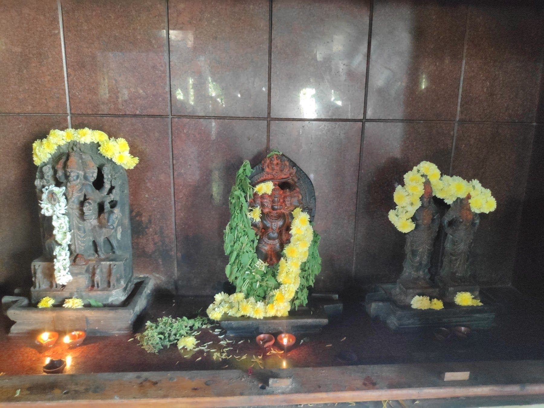 ನಯವಾದ ಗ್ರಾನೈಟ್ ಗೋಡೆಯ ಮುಂದೆ ಹಳದಿ ಹೂಮಾಲೆ ಹಾಕಿದ ಮೂರು ಹಳೆಯ ಶಿಲಾ ಶಿಲ್ಪಗಳು; ಮುಂದೆ ಪುಟ್ಟ ಎಣ್ಣೆ ದೀಪಗಳು